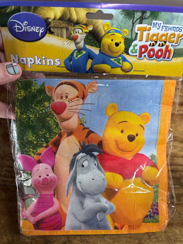 Packaged Disney napkins featuring Tigger, Pooh, Piglet, and Eeyore on a wooden surface.