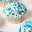 A close-up of a blue and white frosted cupcake topped with blue and white sprinkles, with other cupcakes partially visible in the background on a white surface.