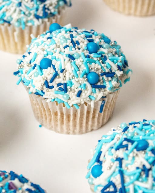 A close-up of a blue and white frosted cupcake topped with blue and white sprinkles, with other cupcakes partially visible in the background on a white surface.