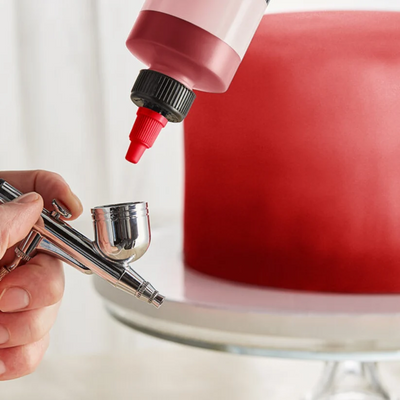 A close-up of a hand holding an airbrush while applying red icing to a large, smooth red cake on a white stand.
