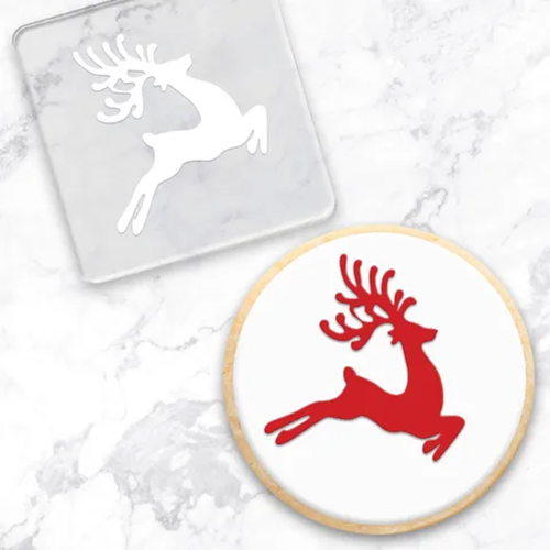 A round white cookie with a red reindeer silhouette, shown next to a clear square acrylic embossing tool with a reindeer silhouette, on a light marble background.