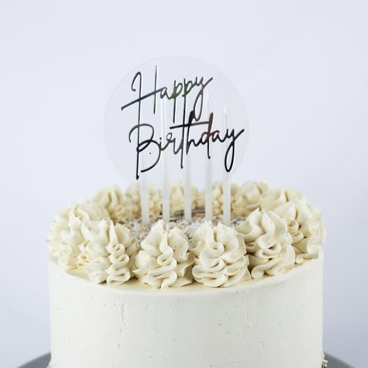 A round white frosted cake with piped cream frosting rosettes around the top edge, featuring a clear circular 'Happy Birthday' cake topper with black script lettering.