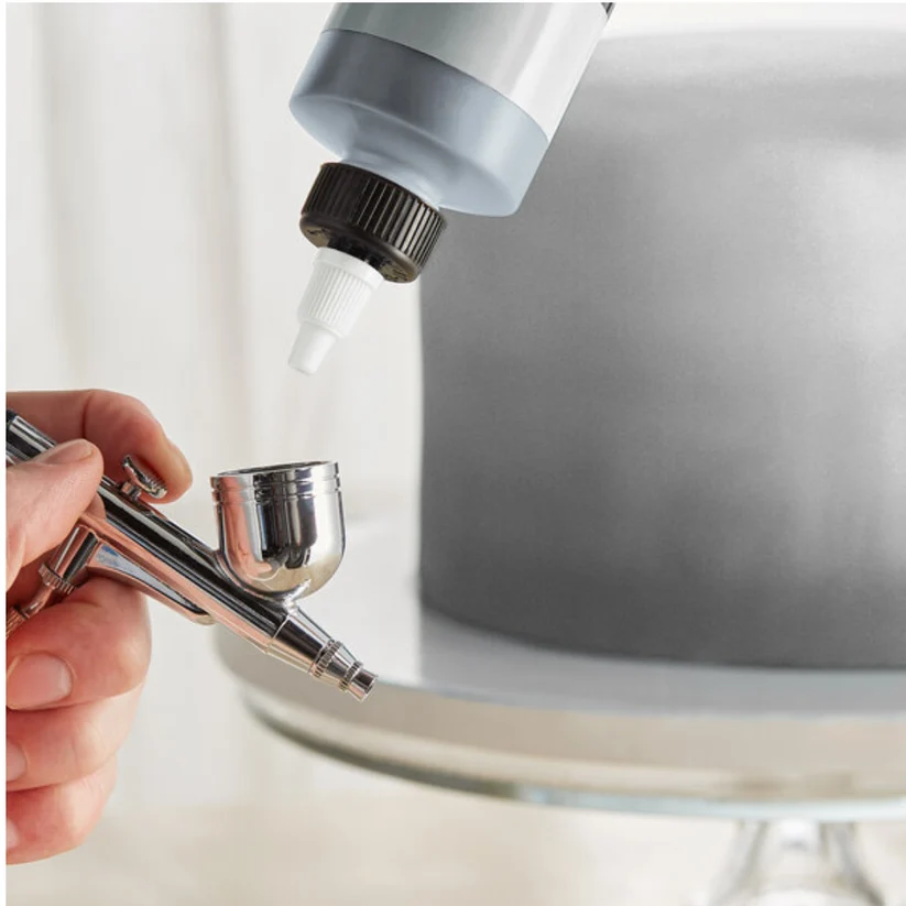 A close-up of a hand holding an airbrush with a bottle of liquid being dispensed above it, with a blurred gray frosted cake on a stand in the background.