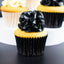 A close-up of a cupcake with glossy black rosette-style frosting on a yellow cake base, in a black pleated liner. Two more cupcakes are softly blurred in the background on a white and dark surface.