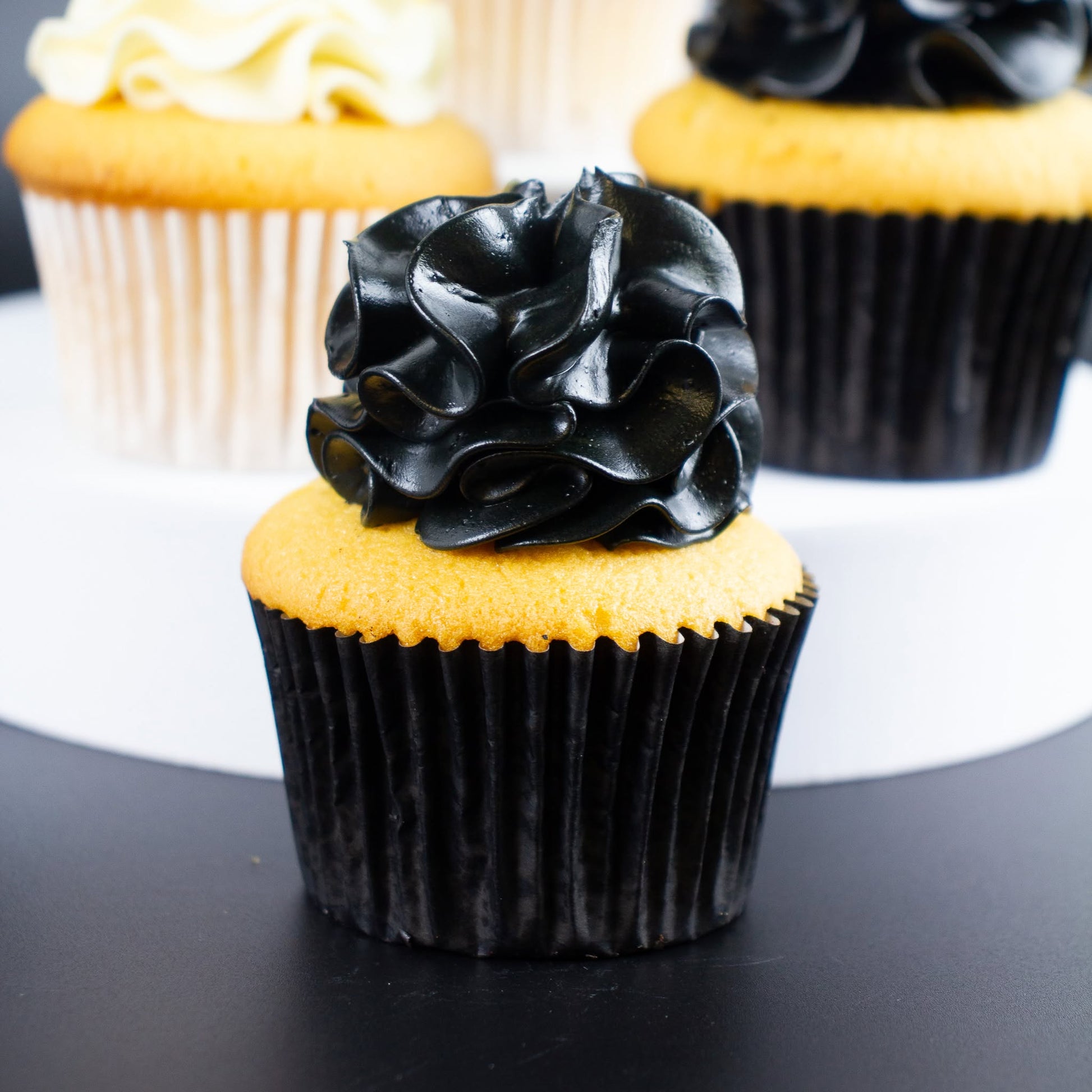 A close-up of a cupcake with glossy black rosette-style frosting on a yellow cake base, in a black pleated liner. Two more cupcakes are softly blurred in the background on a white and dark surface.