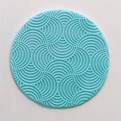A round turquoise blue fondant plaque featuring a repeating scallop or wave pattern, shown on a plain off-white background.