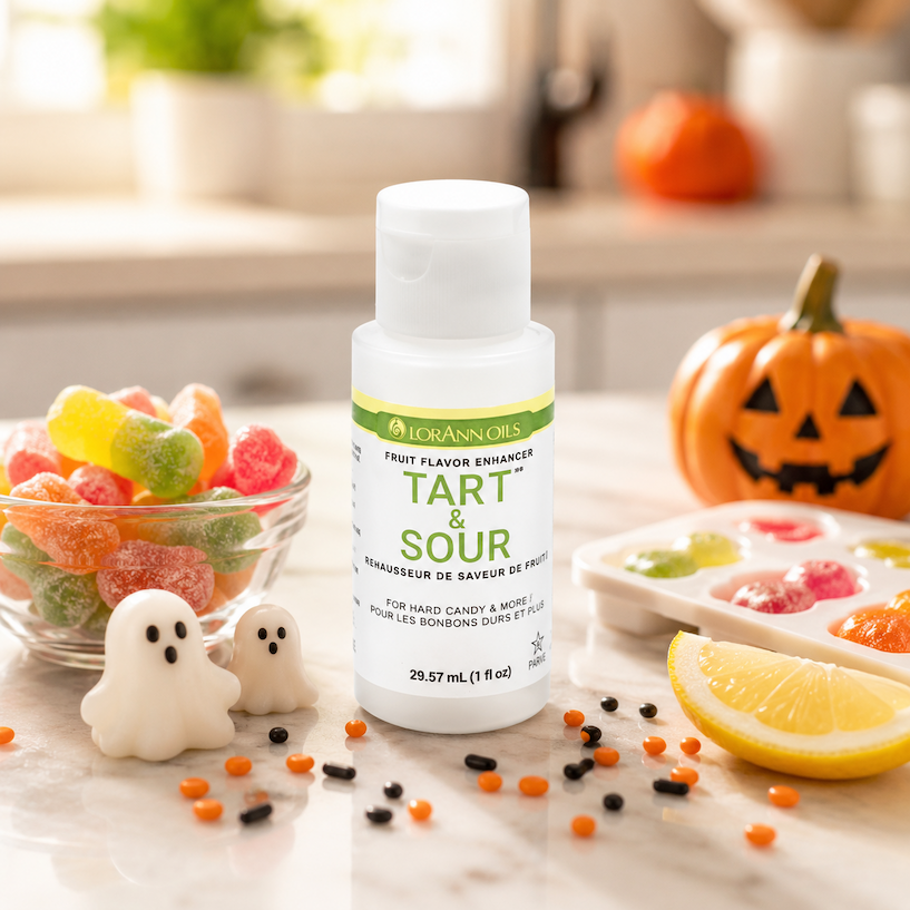 A bottle of Lorann's Tart & Sour flavor enhancer stands on a light countertop, surrounded by Halloween-themed props including a bowl of colorful gummy candy, small ghost decorations, a lemon wedge, scattered orange and black candy pieces, and a jack-o'-lantern in the background.