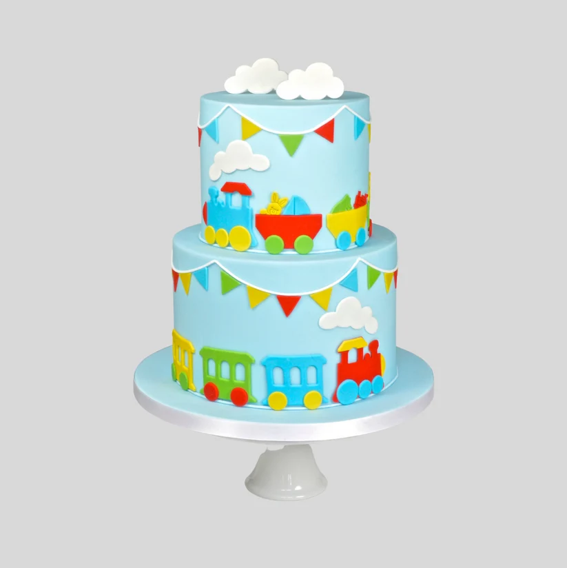 A two-tier round cake decorated with a light blue fondant background, colorful train illustrations, white fondant clouds, and small triangular bunting flags, displayed on a white cake stand against a solid gray background.