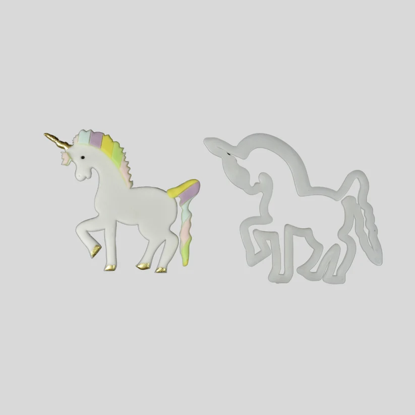 A white unicorn-shaped fondant cut out with a pastel rainbow mane, tail, and gold horn, shown beside a white plastic matching outline cutter on a plain light gray background.