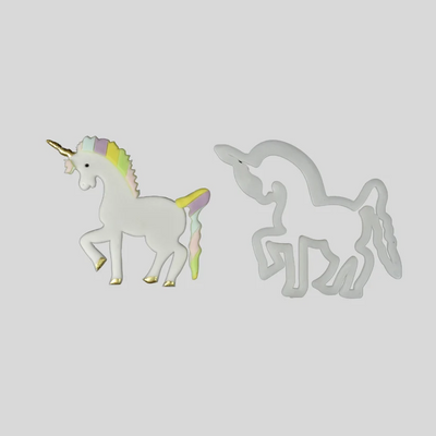 A white unicorn-shaped fondant cut out with a pastel rainbow mane, tail, and gold horn, shown beside a white plastic matching outline cutter on a plain light gray background.