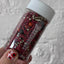 Bottle of sprinkles held by a hand with red nail polish against a white background