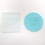 A round blue fondant plaque with a shark outline, shown beside a clear square acrylic embossing tool featuring the same shark design, on a light  surface.