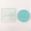 A clear debosser with the word 'Good Luck' embossed on it, placed on a light-colored surface. Next to a blue fondant icing example of use