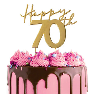 A gold metal cake topper spelling out 'Happy 70th' placed on top of a decorated cake with pink icing and chocolate drips.