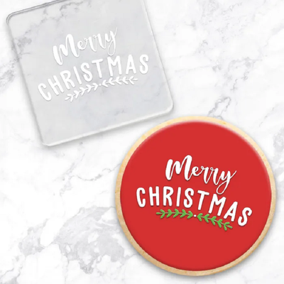 Clear acrylic embosser with Red cookie showcasing how to use it Both read Merry Christmas with a small vine leaf design below the text